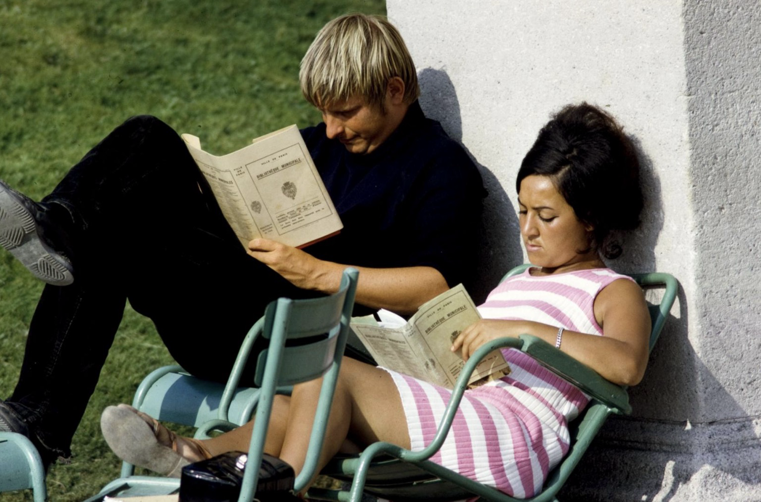 Photographie couleur montrant un couple allongé côte à côte sur des chaises, adossé à un mur du jardin du Luxembourg, en train de lire ensemble un livre